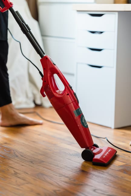 A person using a red corded vacuum cleaner to clean a wooden floor in a room with white furniture. The vacuum is positioned centrally, with the cleaner's handle held by a person whose feet are visible, standing barefoot on the wooden surface. To the right, a white chest of drawers with multiple small drawers is visible, illuminated by natural light. The room appears tidy and well-maintained, reflecting a domestic environment. This scene demonstrates surface cleaning and deep cleaning practices that promote hygiene, aligning with the services provided by Carpet Cleaning Hackney for professional residential cleaning. The focus on the vacuum and clean wooden flooring emphasizes the importance of thorough cleaning and sanitisation in home maintenance.