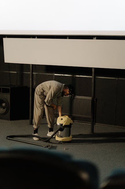 A person wearing a beige jumpsuit is performing industrial surface cleaning in a commercial space, using a yellow and black vacuum cleaner placed on the carpeted floor. The individual is bent over slightly, maneuvering the cleaning equipment towards the carpet area beneath a large, white, wall-mounted panel or acoustic ceiling feature. The room features dark-colored walls with metallic accents, and natural light is coming from an unseen source, highlighting the cleanliness and maintenance process. Visible equipment and the professional cleaning environment reflect deep cleaning and sanitisation practices typical of commercial carpet and surface maintenance, consistent with the services offered by Carpet Cleaning Hackney in the Dalston E8 area.