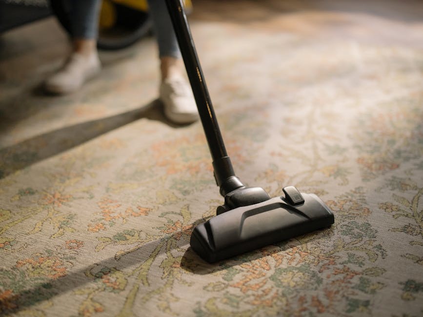A person using a red corded vacuum cleaner to clean a wooden floor in a room with white furniture. The vacuum is positioned centrally, with the cleaner's handle held by a person whose feet are visible, standing barefoot on the wooden surface. To the right, a white chest of drawers with multiple small drawers is visible, illuminated by natural light. The room appears tidy and well-maintained, reflecting a domestic environment. This scene demonstrates surface cleaning and deep cleaning practices that promote hygiene, aligning with the services provided by Carpet Cleaning Hackney for professional residential cleaning. The focus on the vacuum and clean wooden flooring emphasizes the importance of thorough cleaning and sanitisation in home maintenance.