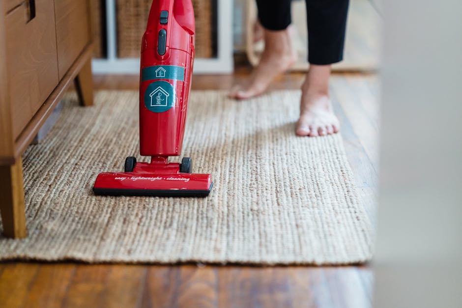 A professional cleaner is performing deep surface cleaning in a modern, well-lit room with a dark tile floor and black wall panels. The cleaner, dressed in a light grey jumpsuit, is bent over focused 