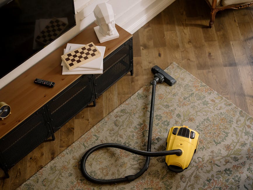 A yellow canister vacuum cleaner with a flexible black hose and attachment is positioned on an ornate, multicolored area rug in a living room. The room has wooden flooring and features a black metal and wood media unit against a white wall, on top of which hang a remote control, a chessboard, and a white sculptural bust. The lighting is natural, casting soft shadows across the surfaces, and the overall scene depicts a clean, well-maintained interior space prepared for deep surface cleaning, with visible attention to hygiene and maintenance, as part of a professional domestic cleaning service by Carpet Cleaning Hackney.