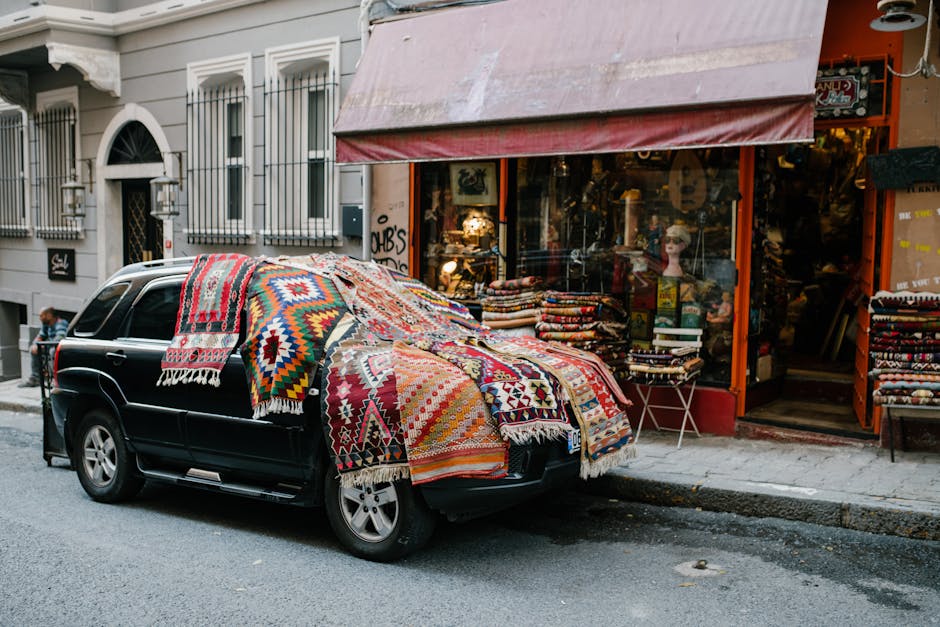 An indoor display of traditional rugs with intricate geometric and floral patterns, hanging on a wooden lattice frame. Several rugs with detailed motifs are draped over benches and shelves, showcasing