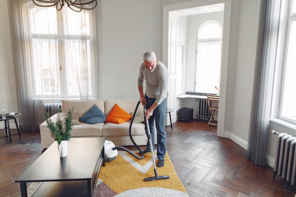 A middle-aged man with grey hair and a beard is engaged in deep surface cleaning of a living room in a residential home, using a vacuum cleaner. He is wearing a light grey long-sleeve shirt and blue jeans, standing on a wooden floor with a yellow geometric patterned rug. The room features a white sofa with assorted throw pillows in blue and orange, next to a black side table with a glass of water. Large windows with sheer white curtains allow natural light to fill the space, highlighting the pristine, dust-free floor and well-maintained white walls. Visible accessories include a potted plant on the coffee table and a corner desk with a wooden chair, complemented by a radiator beneath the windows. The scene exemplifies professional domestic cleaning and surface maintenance, with Carpet Cleaning Hackney ensuring thorough hygiene and surface care in this inviting, tidy living room.