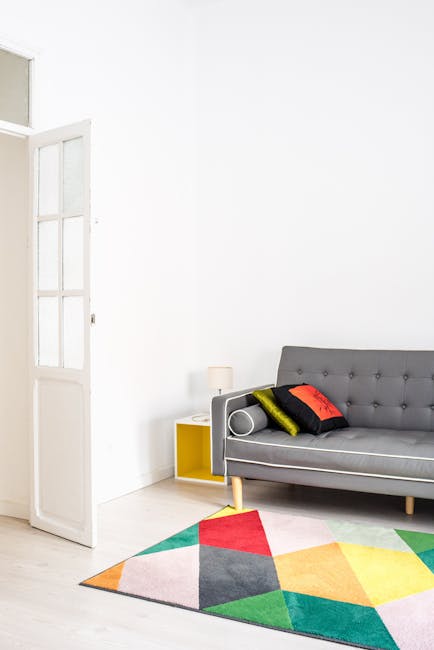 A modern living room featuring a light-colored hardwood floor with a multicolored geometric rug in shades of pink, green, yellow, red, gray, and black. Against a white wall, there is a gray upholstered sofa with a tufted back and wooden legs, decorated with three pillows in black, yellow, and red. To the left, a small yellow side table holds a white lamp with a cylindrical shade. Partially visible on the left side of the image is a white door with glass panels, slightly ajar, allowing natural light to fill the space. The room appears tidy and well-maintained, highlighting a scene of domestic surface cleaning and sanitisation, in line with services offered by Carpet Cleaning Hackney, situated near Hackney Wick station.