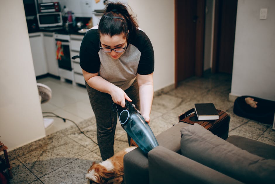 A middle-aged man with grey hair and a beard is engaged in deep surface cleaning of a living room in a residential home, using a vacuum cleaner. He is wearing a light grey long-sleeve shirt and blue jeans, standing on a wooden floor with a yellow geometric patterned rug. The room features a white sofa with assorted throw pillows in blue and orange, next to a black side table with a glass of water. Large windows with sheer white curtains allow natural light to fill the space, highlighting the pristine, dust-free floor and well-maintained white walls. Visible accessories include a potted plant on the coffee table and a corner desk with a wooden chair, complemented by a radiator beneath the windows. The scene exemplifies professional domestic cleaning and surface maintenance, with Carpet Cleaning Hackney ensuring thorough hygiene and surface care in this inviting, tidy living room.
